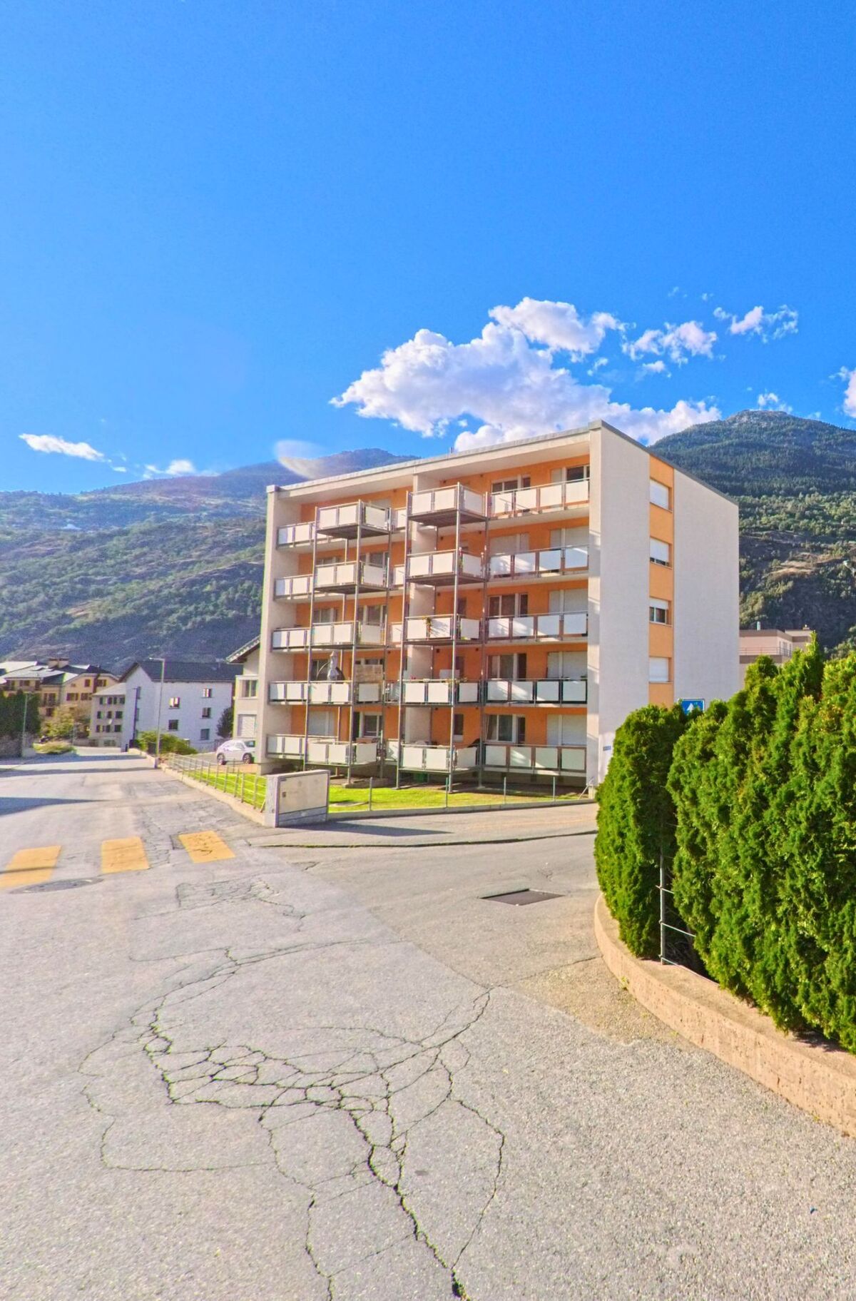 Ein mehrstöckiges Wohnhaus mit Balkonen erhebt sich in Brig-Glis (3902, Kanton Wallis) vor der Kulisse bewaldeter Berghaenge.