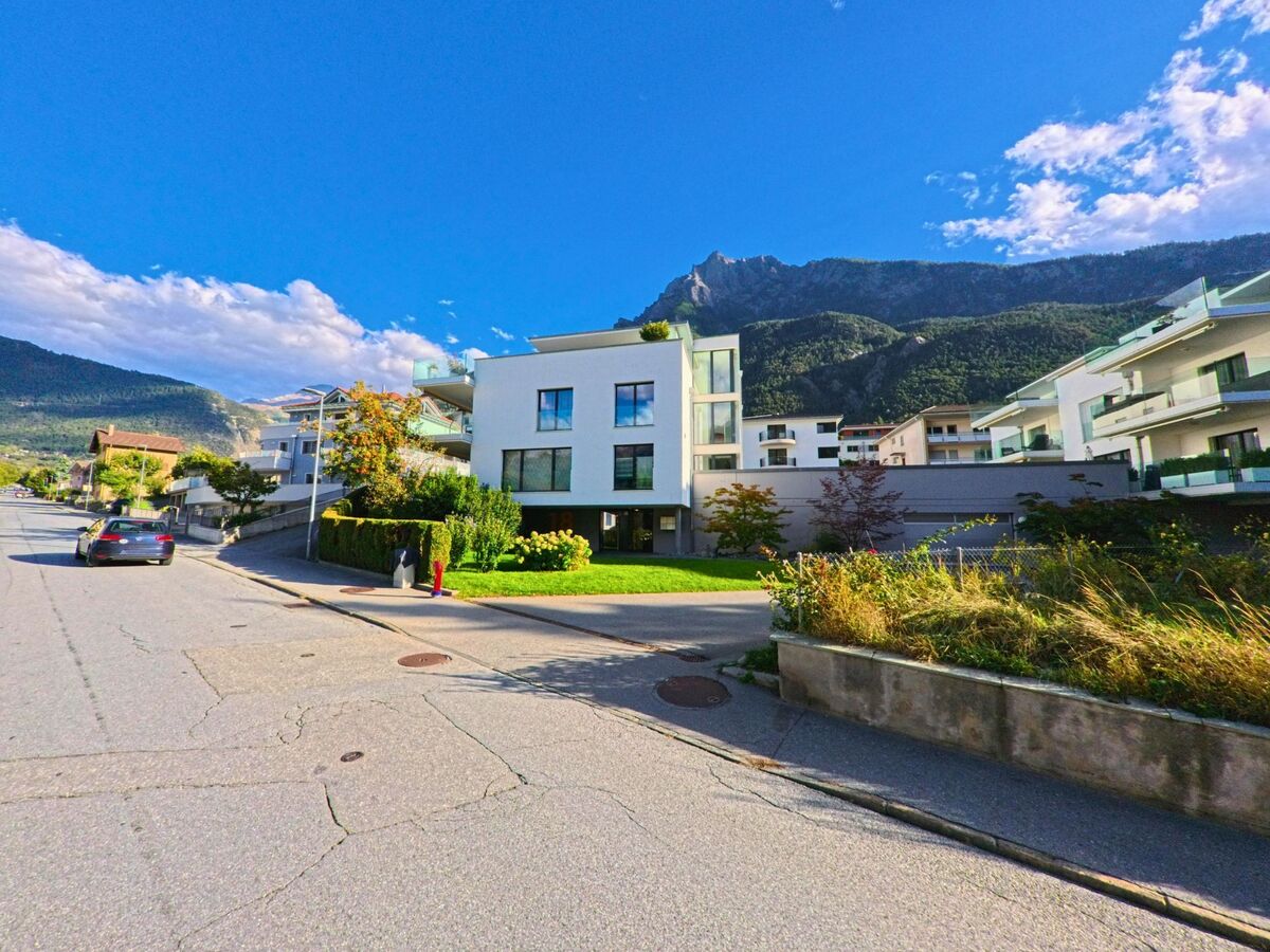 Eine moderne Wohnsiedlung in Brig-Glis (3902, Kanton Wallis) liegt am Fuss der bewaldeten Berghaenge unter blauem Himmel.