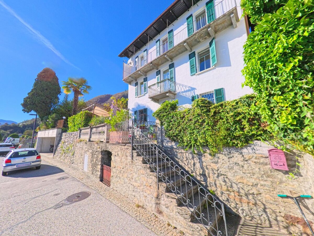 Ein weisses Haus mit grünen Fensterläden und Balkon steht an einer sonnigen Strasse in Brione sopra Minusio (6645, Kanton Tessin).
