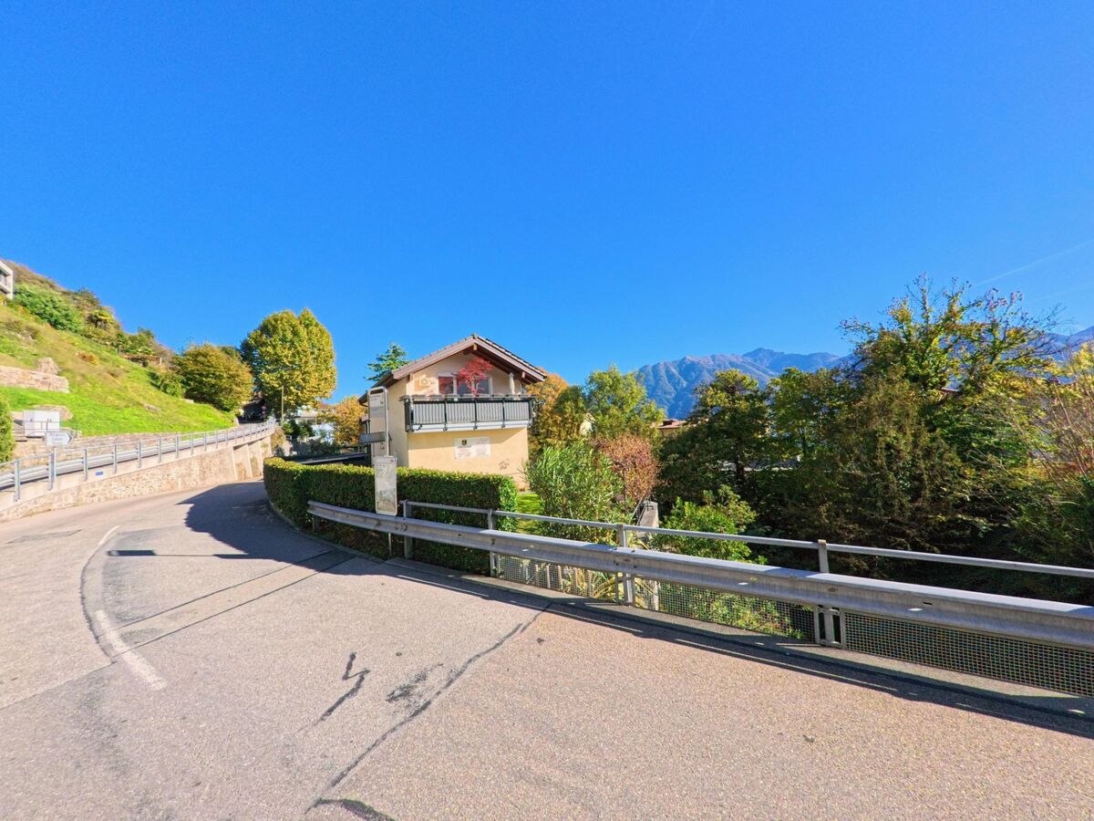 Eine kurvige Strasse führt durch Brione sopra Minusio (6645, Kanton Tessin) vorbei an einem Haus mit Balkon und gepflegter Hecke.