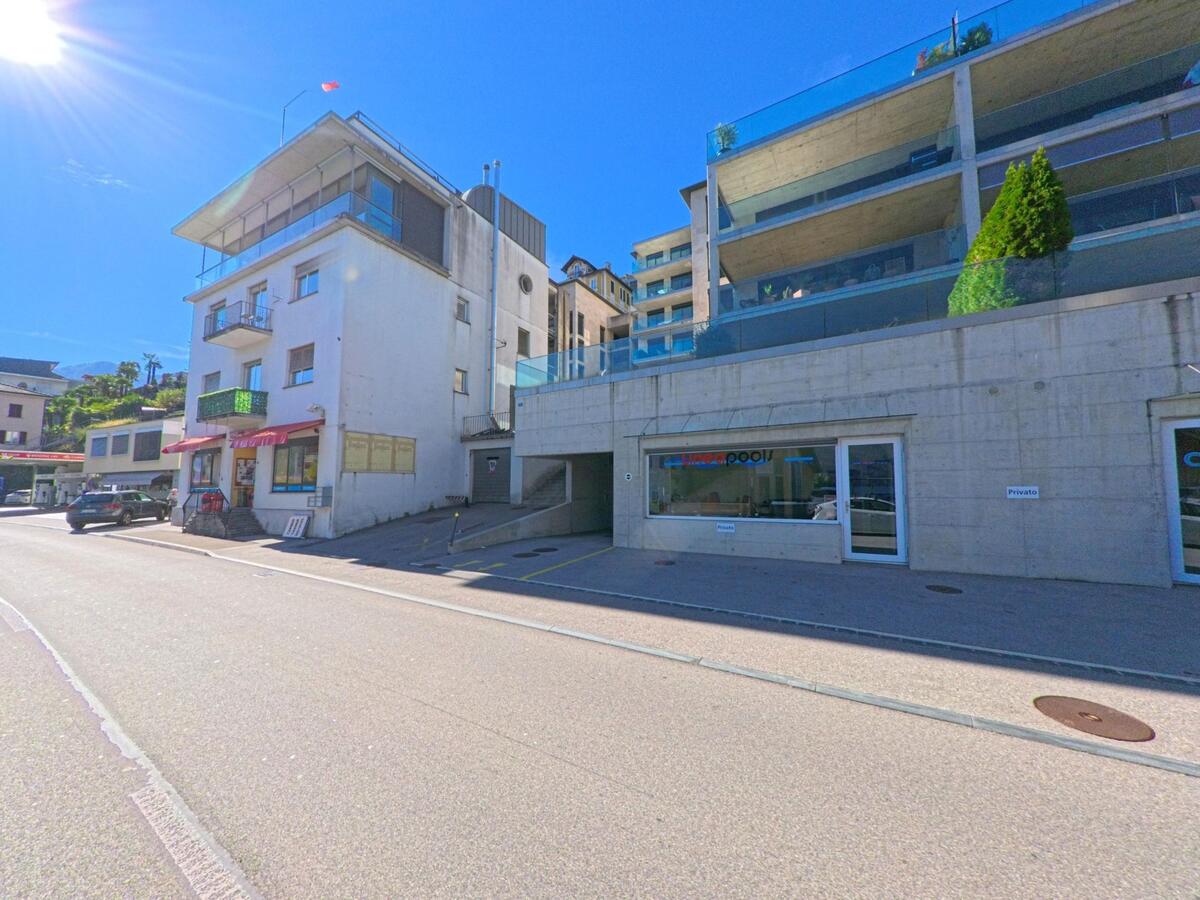 In Brissago (6614, Kanton Tessin) stehen moderne und ältere Gebäude nebeneinander an einer sonnigen Strasse mit Geschäften und Parkplätzen.