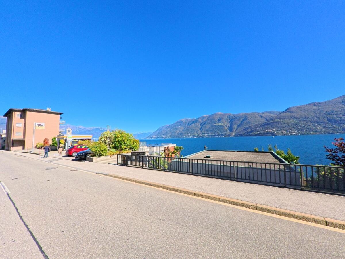 In Brissago (6614, Kanton Tessin) führt eine Strasse mit Geländer am Lago Maggiore entlang, mit Blick auf die Berge am gegenüberliegenden Ufer