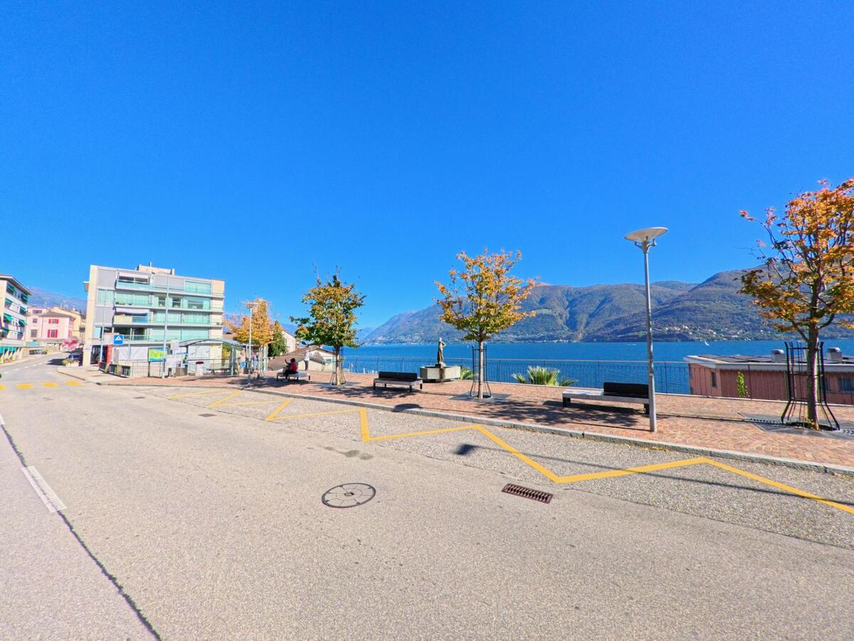 In Brissago (6614, Kanton Tessin) liegt eine Uferpromenade mit Bänken und Bäumen direkt am Lago Maggiore unter klarem blauem Himmel.