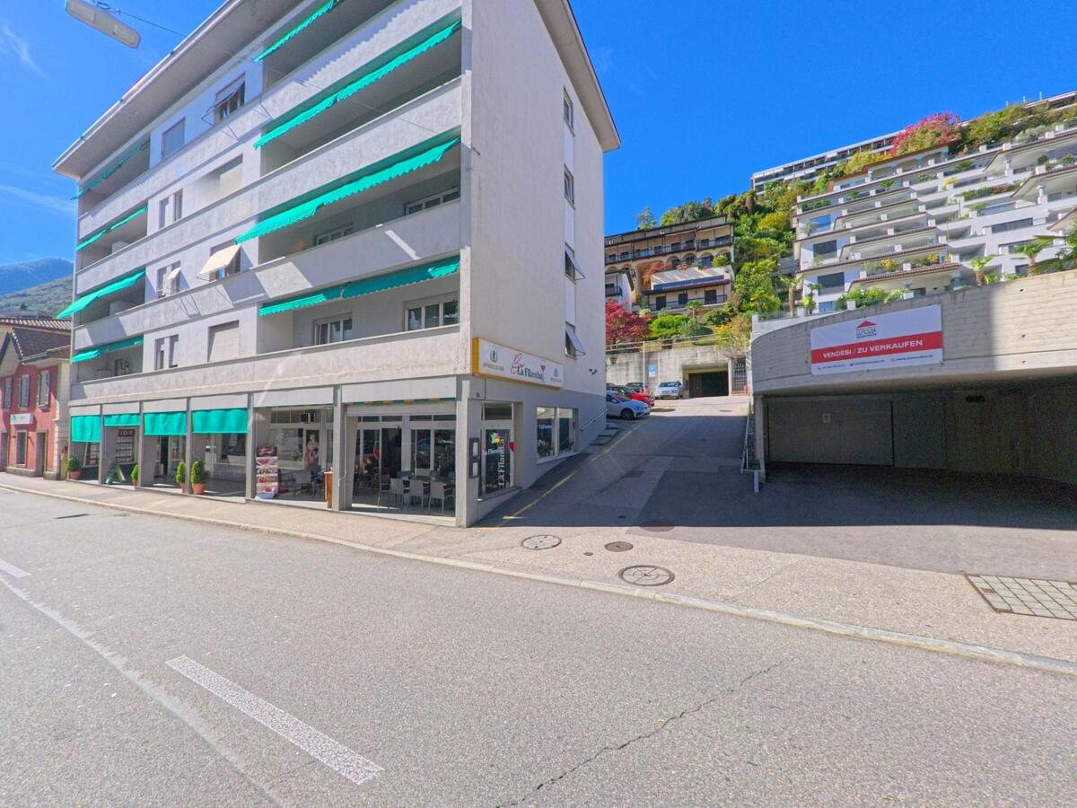In Brissago (6614, Kanton Tessin) steht ein Wohnhaus mit grünen Markisen und einem Café an der Strasse, daneben führt eine Rampe bergauf.