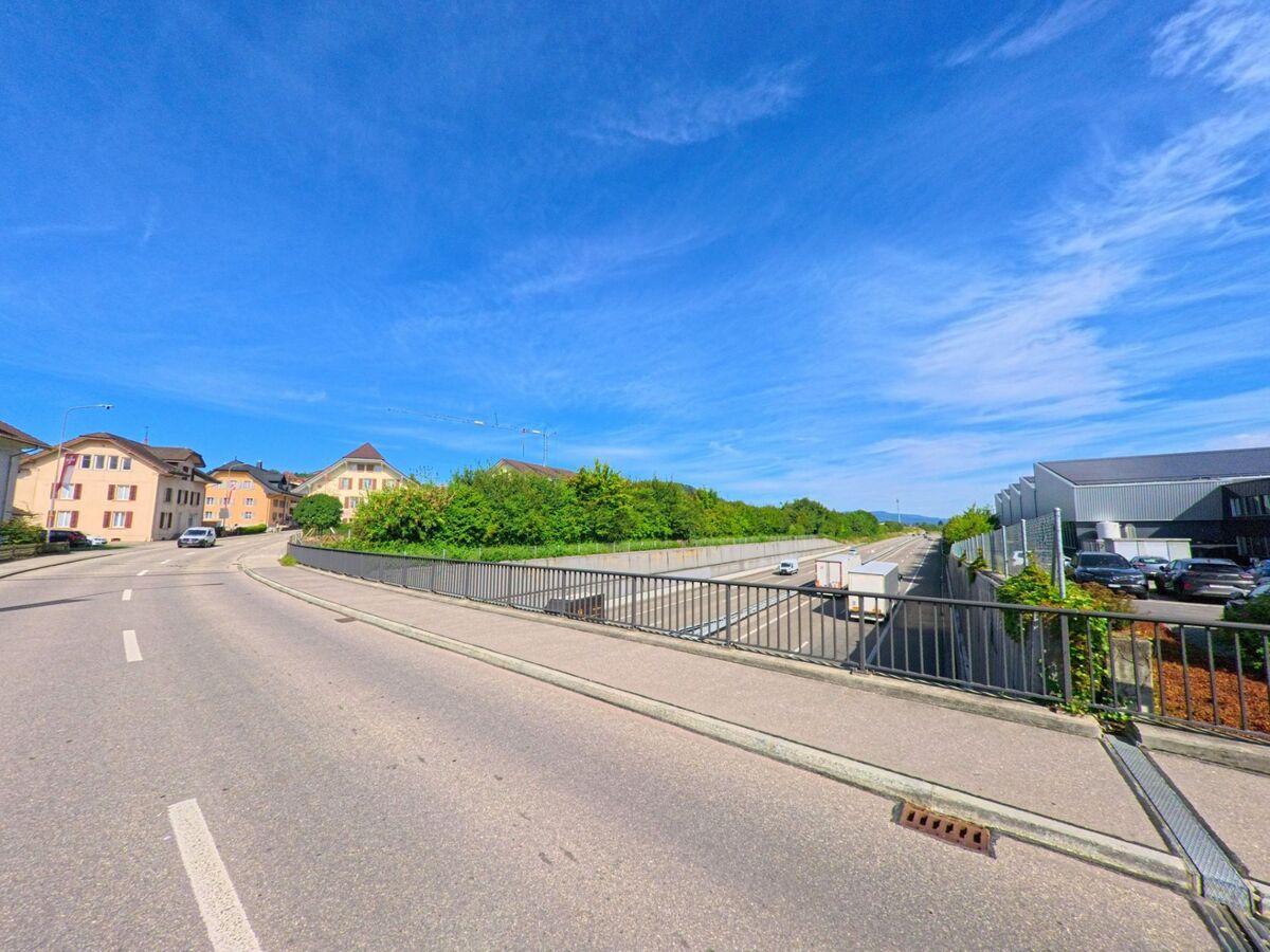  In Brittnau (4805, Kanton Aargau) fuehrt eine Bruecke mit Geländer ueber die Autobahn, waehrend auf der einen Seite Wohnhaeuser und auf der anderen ein Gewerbebau liegen.