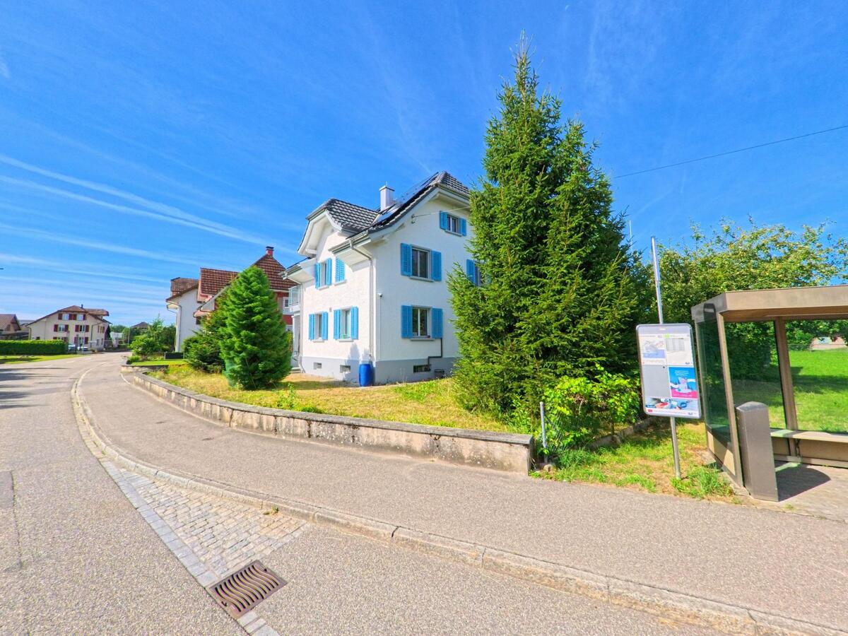 In Brittnau (4805, Kanton Aargau) steht ein helles Wohnhaus mit blauen Fensterlaeden neben einer kleinen Bushaltestelle an einer ruhigen Quartierstrasse.