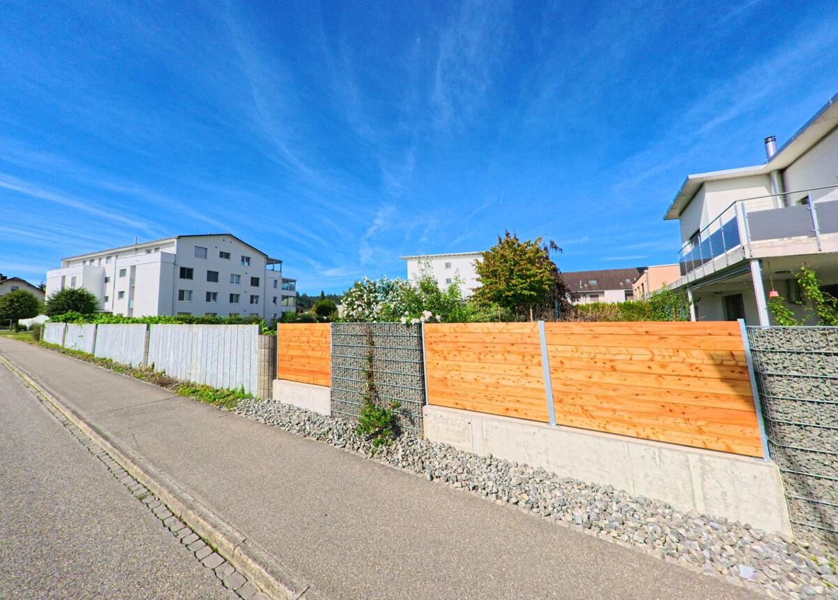  In Brittnau (4805, Kanton Aargau) stehen moderne Mehrfamilienhaeuser hinter einer Kombination aus Holz- und Gabionenzaun entlang eines ruhigen Fusswegs.