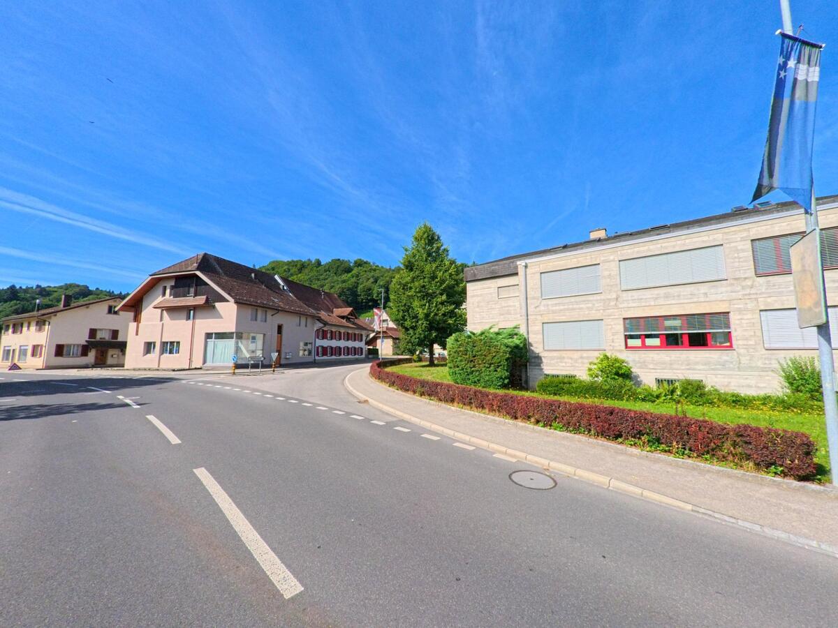 In Brittnau (4805, Kanton Aargau) verlaeuft eine Strasse zwischen einem Schulgebaeude mit grauer Fassade und traditionellen Wohnhaeusern mit Fensterlaeden.