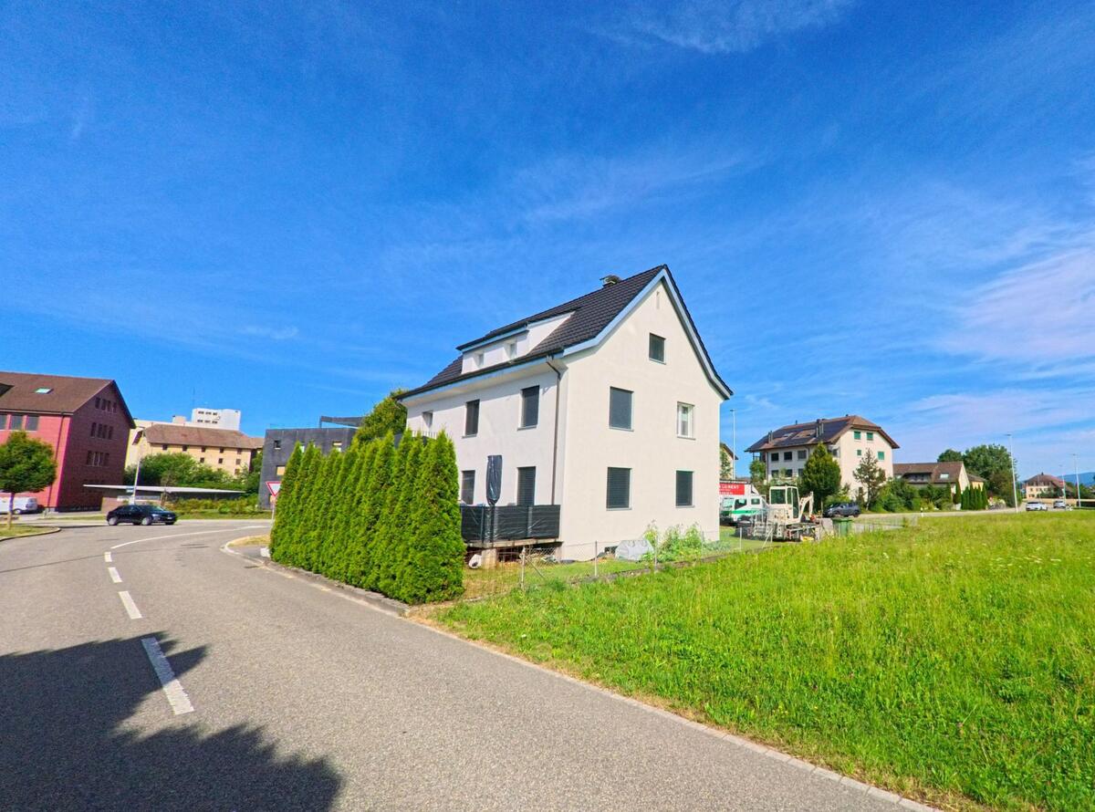 In Brittnau (4805, Kanton Aargau) steht ein schlichtes Wohnhaus mit dunklen Fensterlaeden an einer Kurve, umgeben von Wiesen und weiteren Siedlungsbauten.