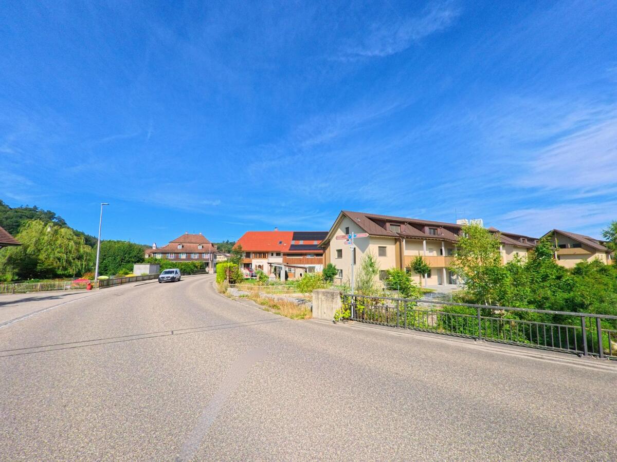 In Brittnau (4805, Kanton Aargau) verläuft eine leicht ansteigende Strasse mit Einfamilienhaeusern und Mehrfamilienbauten, flankiert von einem schlichten Brueckengeländer.