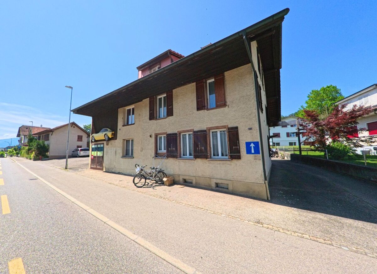 In Brügg (2555, Kanton Bern) führt eine Straße an einem traditionellen Gebäude entlang, das durch ein auffälliges, an der Wand montiertes Auto gekennzeichnet ist.