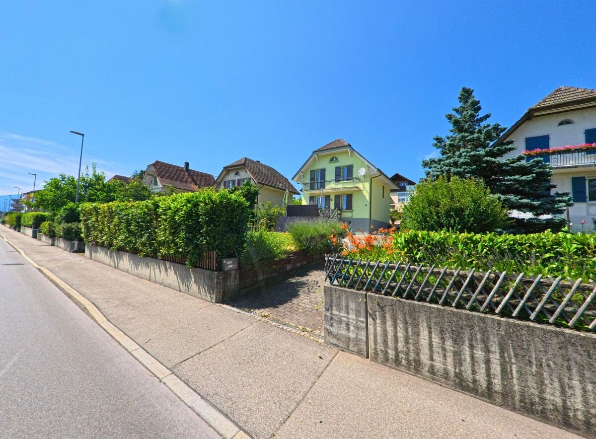 In Brügg (2555, Kanton Bern) führt eine Straße an gepflegten Gärten und traditionellen Häusern entlang, während Bäume und Blumen das Bild zieren.