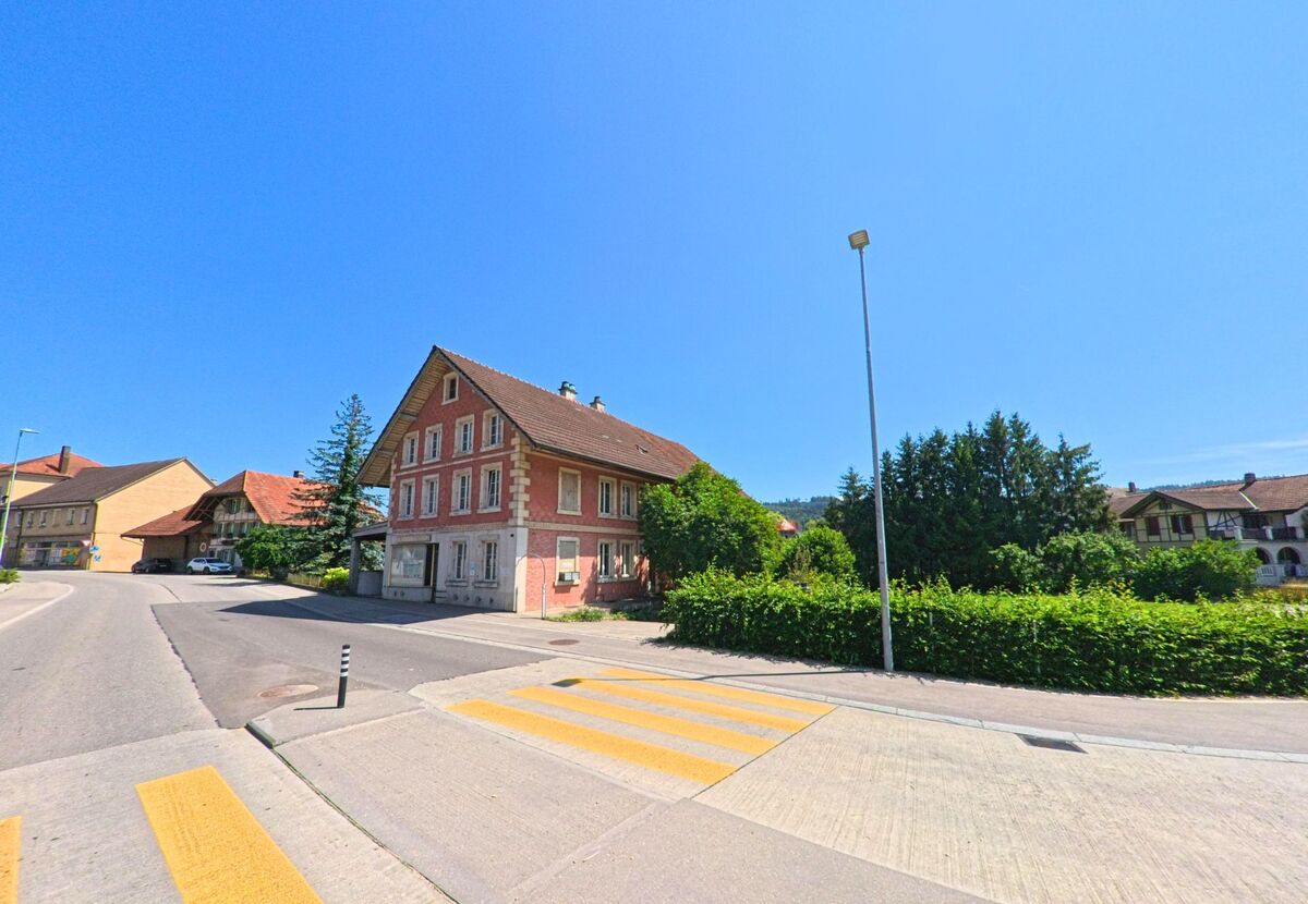 In Brügg (2555, Kanton Bern) führt eine Straße an einem traditionellen, rot gestrichenen Gebäude entlang, während im Hintergrund weitere Häuser und Bäume sichtbar sind.