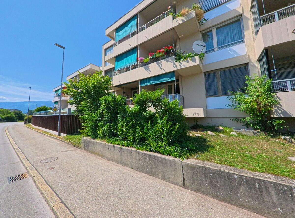 In Brügg (2555, Kanton Bern) verläuft eine Straße an einem modernen Mehrfamilienhaus entlang, das mit Balkonen und grünen Pflanzen geschmückt ist.