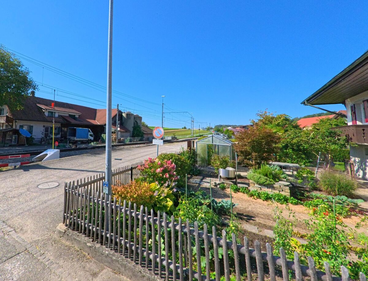In Brüttelen (3237, Kanton Bern) führt ein Bahnübergang mit Schranken an Wohn- und Wirtschaftsgebäuden sowie einem kleinen Garten vorbei.