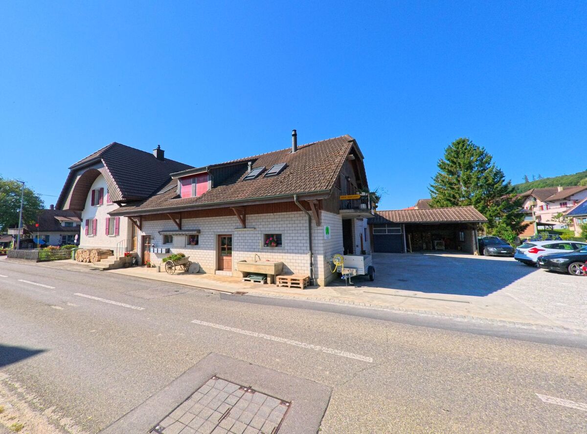 Entlang der Hauptstrasse in Brüttelen (3237, Kanton Bern) steht ein Bauernhaus mit weisser Fassade und roten Fensterläden neben einem Nebengebäude.
