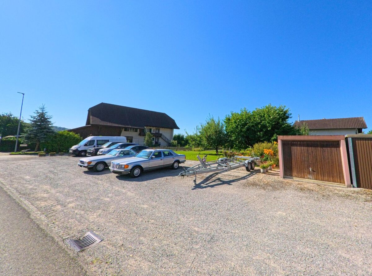 Am Dorfrand von Brüttelen (3237, Kanton Bern) stehen mehrere geparkte Fahrzeuge vor Garagen und einem Wohnhaus mit dunklem Dach.