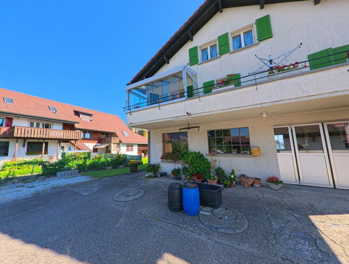 In Brüttelen (3237, Kanton Bern) liegt ein Wohnhaus mit Balkon und grünen Fensterläden gegenüber einem länglichen Gebäude mit Holzverkleidung.