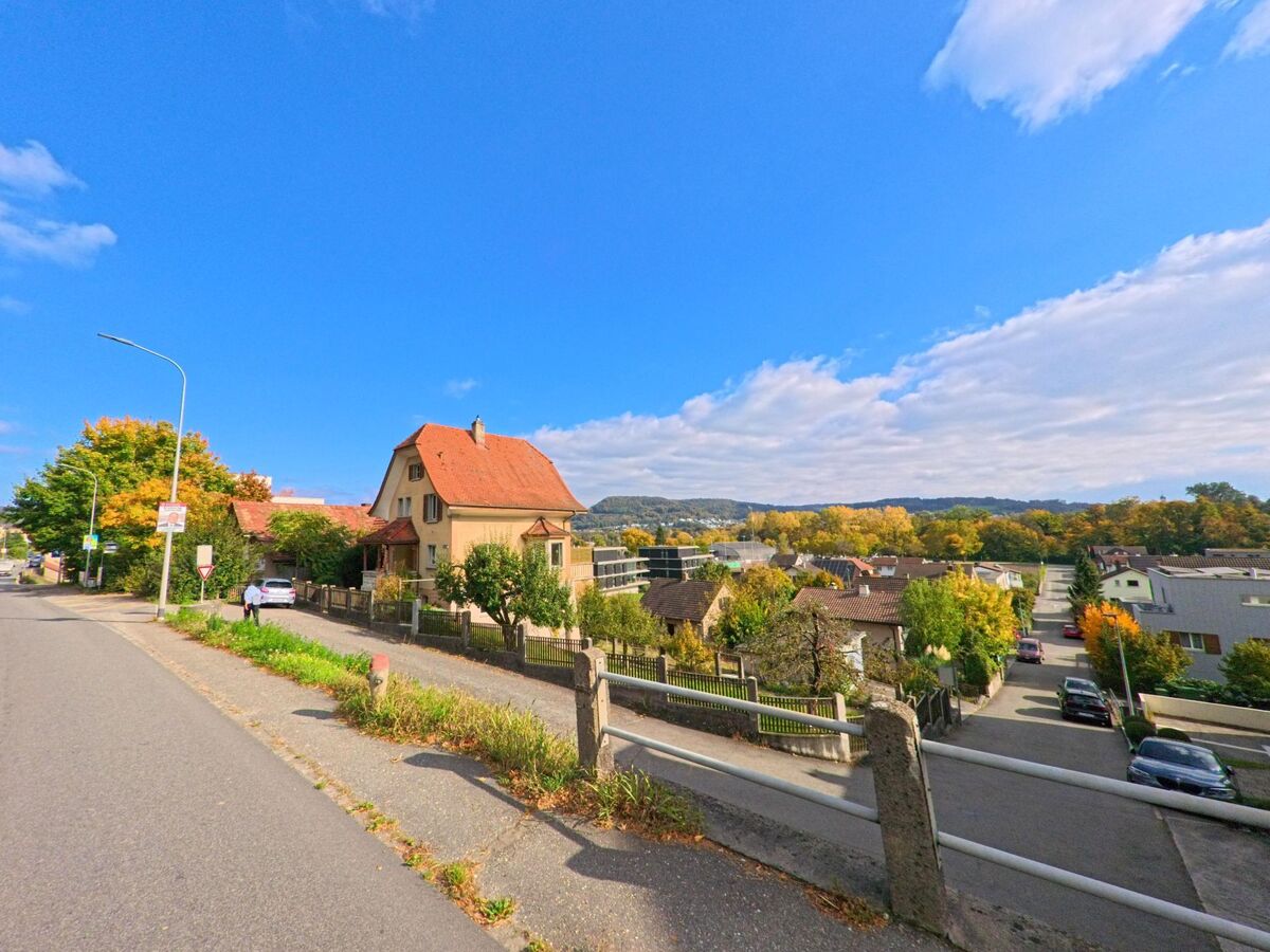 In Brugg (5200, Kanton Aargau) erstreckt sich eine Wohnsiedlung unter blauem Himmel, waehrend am Strassenrand ein Haus mit rotem Dach den Blick ueber die Umgebung von [ORT] begleitet.