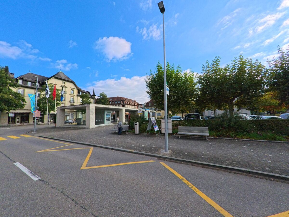 Am Bahnhofplatz in Brugg (5222, Kanton Aargau) befinden sich moderne Pavillons und Sitzbänke, umgeben von Bäumen und historischen Gebäuden im Hintergrund.