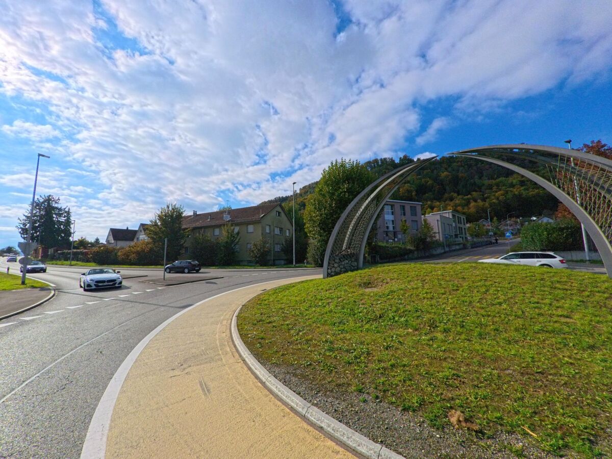 Am Stadtrand von Brugg (5235, Kanton Aargau) führt ein Kreisverkehr mit moderner Bogenkonstruktion an Wohnhäusern und bewaldeten Hängen vorbei.