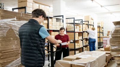 Mitarbeitende arbeiten zwischen Regalen mit Kartons in einem Lagerraum in Zürich (8045, Kanton Zürich) und bereiten Materialien gemeinsam vor.