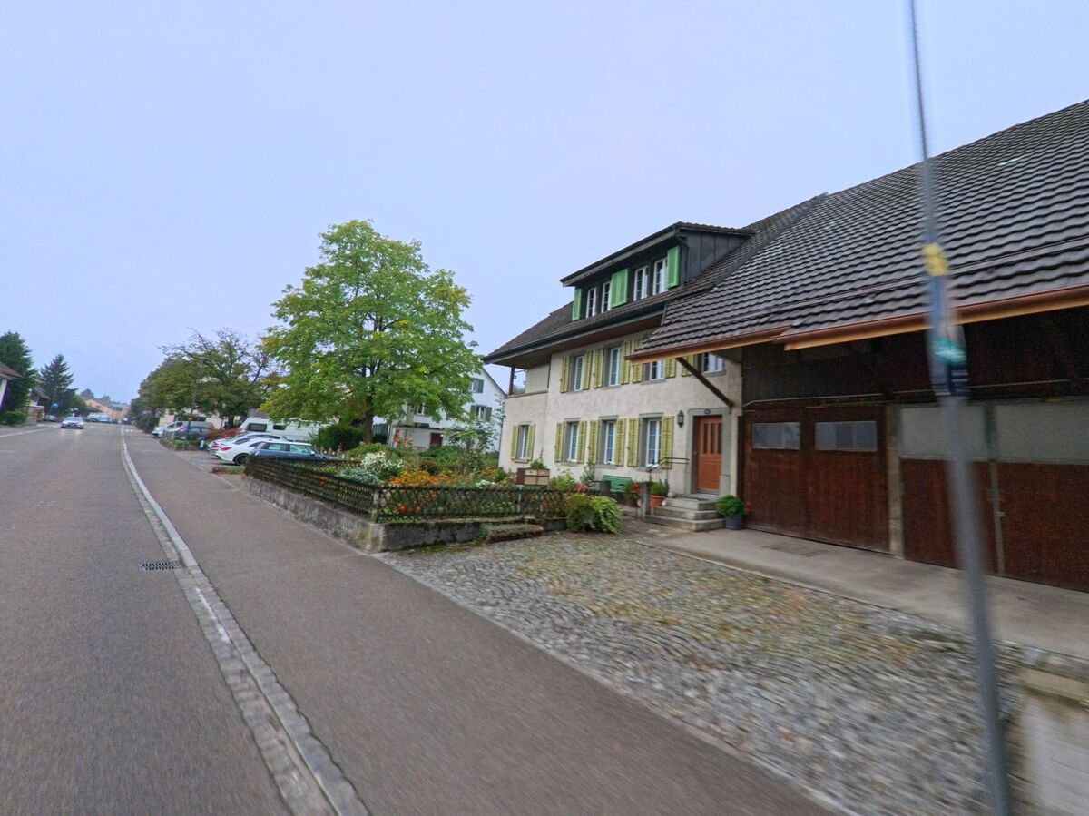 Ein traditionelles Wohnhaus mit grünen Fensterläden und kleinem Blumengarten steht in Brunegg (5505, Kanton Aargau) an einer breiten Dorfstrasse.