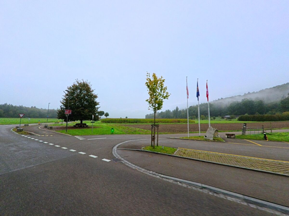 Ein Kreisverkehr in Brunegg (5505, Kanton Aargau) führt an Feldern und Fahnenmasten vorbei, während Nebel über den bewaldeten Hügeln liegt.