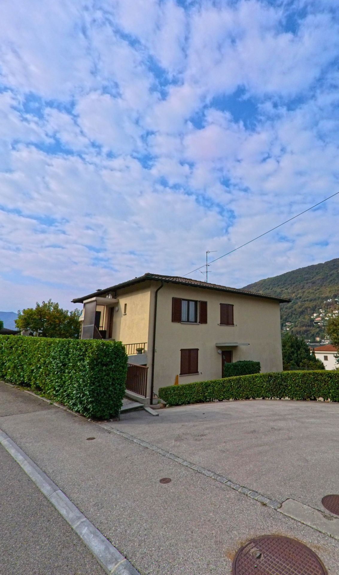 Ein helles Einfamilienhaus mit braunen Fensterläden steht in Brusino Arsizio (6827, Kanton Tessin) an einer ruhigen Seitenstrasse mit gepflegten Hecken.