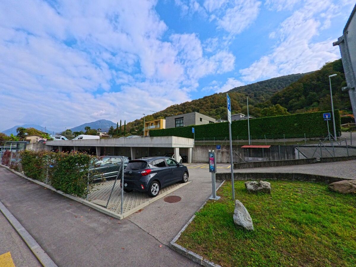 Ein kleiner Parkplatz mit mehreren Autos liegt am Ortsrand von Brusino Arsizio (6827, Kanton Tessin) vor modernen Gebäuden am Fuss bewaldeter Hügel.