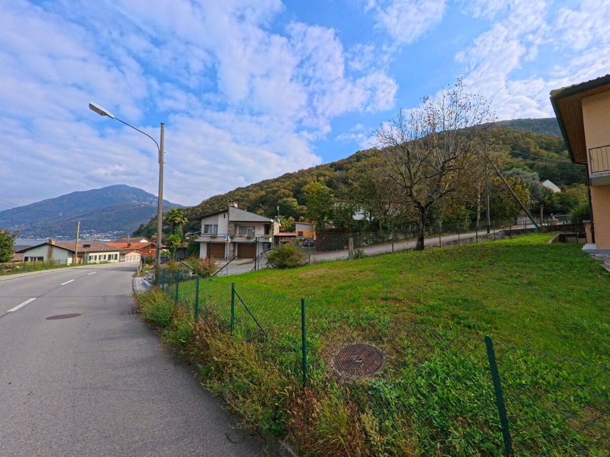Eine leicht ansteigende Strasse führt in Brusino Arsizio (6827, Kanton Tessin) vorbei an Einfamilienhäusern und grünen Gärten mit Blick auf die umliegenden Berge.