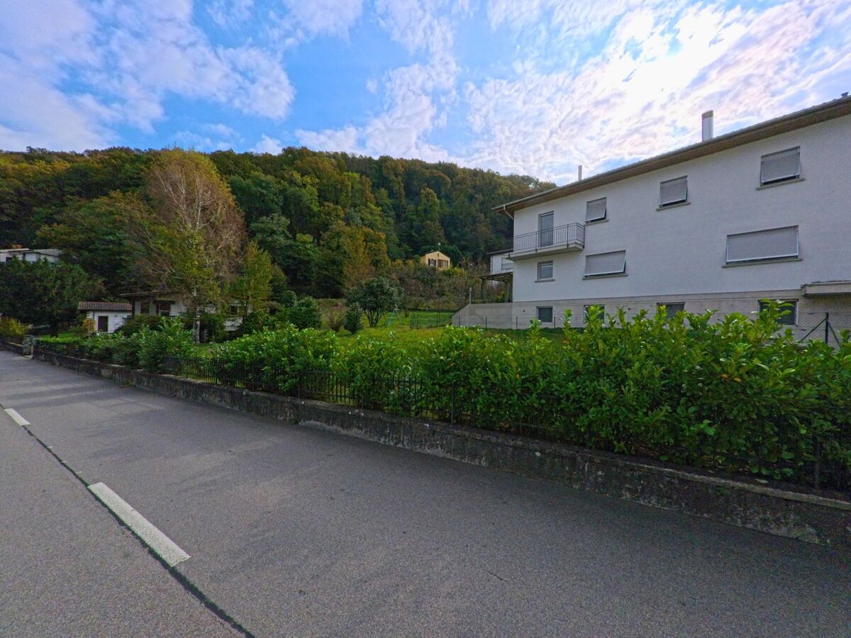 An einer ruhigen Strasse in Brusino Arsizio (6827, Kanton Tessin) stehen Wohnhäuser mit Blick auf grüne Gärten und den bewaldeten Hang im Hintergrund.