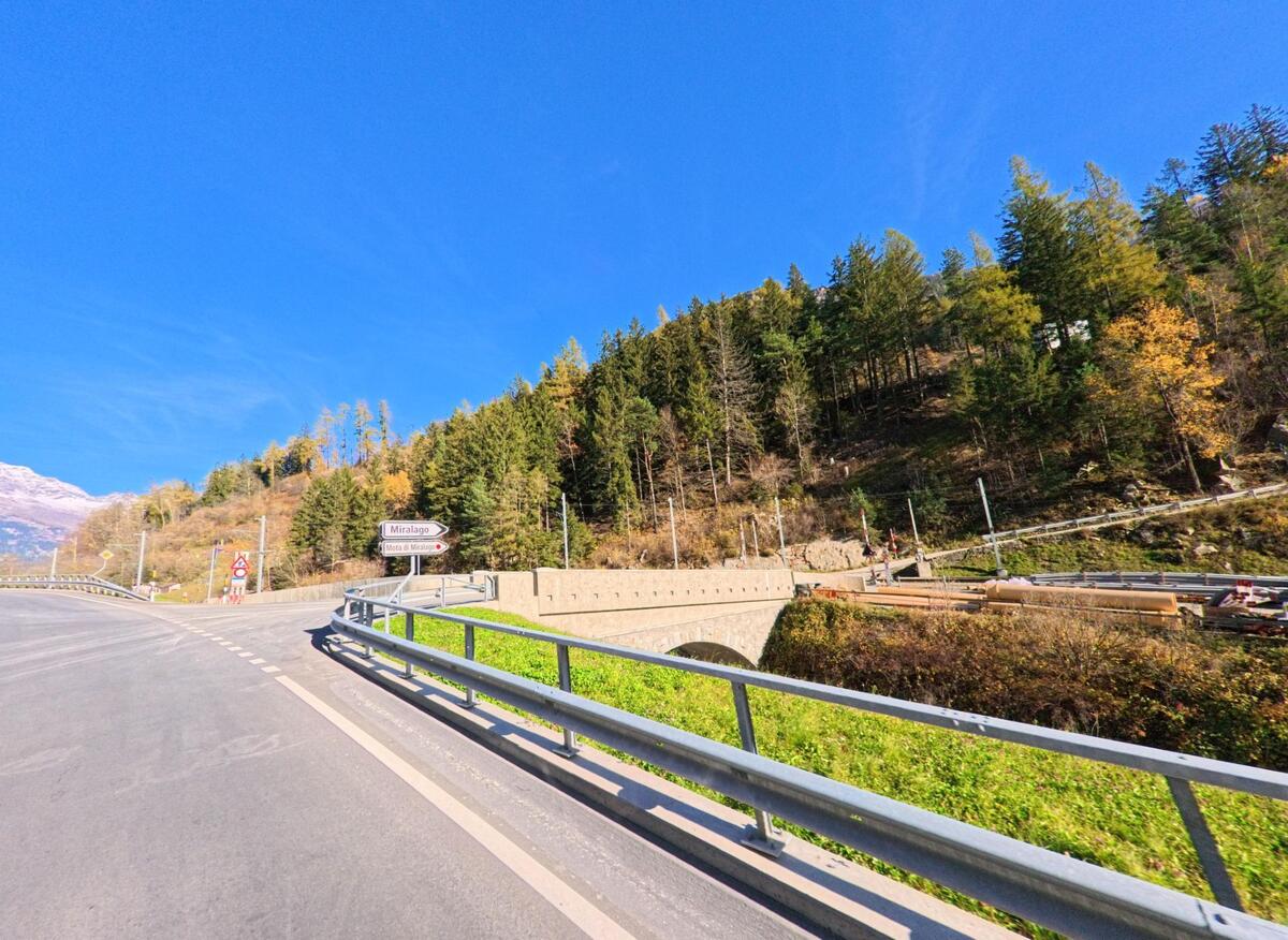 Eine kurvige Strasse mit Leitplanke fuehrt in Brusio (7743, Kanton Graubuenden) an einem bewaldeten Hang vorbei und ueberquert eine kleine Bruecke bei klarem Himmel.