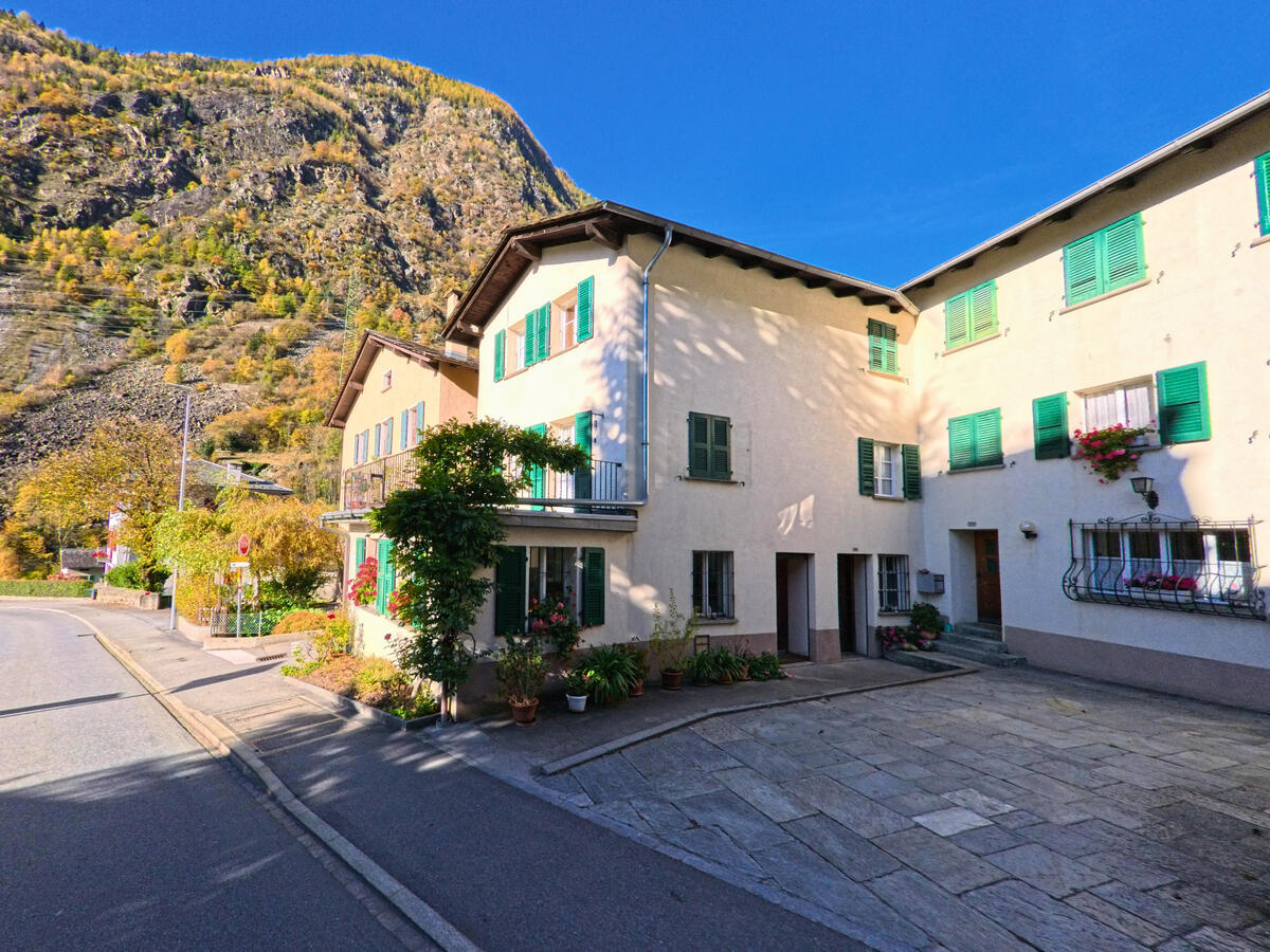 In Brusio (7743, Kanton Graubünden) stehen entlang einer schmalen Dorfstrasse mehrere Wohnhaeuser mit Blick auf die umliegenden Berghaenge.