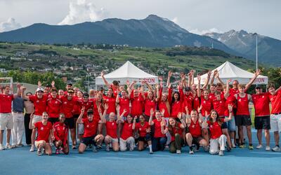In Aarau (5000, Kanton Aargau) posiert eine grosse Gruppe von BTV Aarau Athletics in roten Shirts auf einer Leichtathletikanlage mit erhobenen Armen vor einer Bergkulisse.
