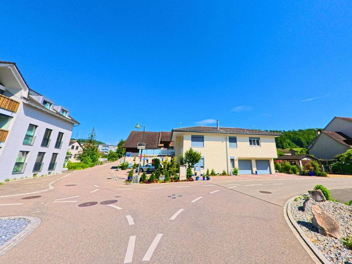 In Bubendorf (4416, Kanton Baselland) oeffnet sich eine verkehrsberuhigte Wohnstrasse zu einem kleinen Platz mit moderner Bebauung und Blick auf den nahen Hang.