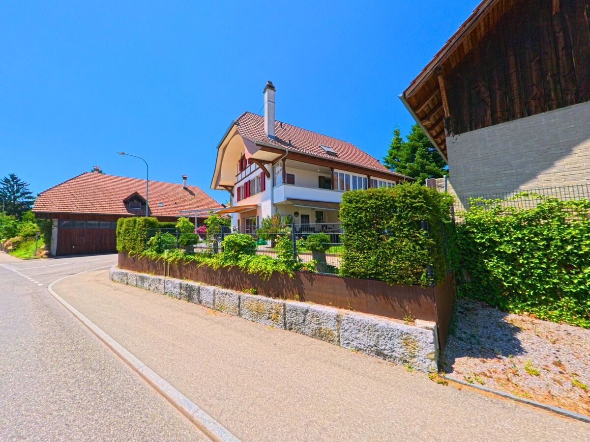 Ein Wohnhaus mit roten Fensterläden und Balkon steht in Buchegg (4576, Kanton Solothurn) an einer ruhigen Strassenecke, umgeben von gepflegten Hecken und Nachbargebäuden.