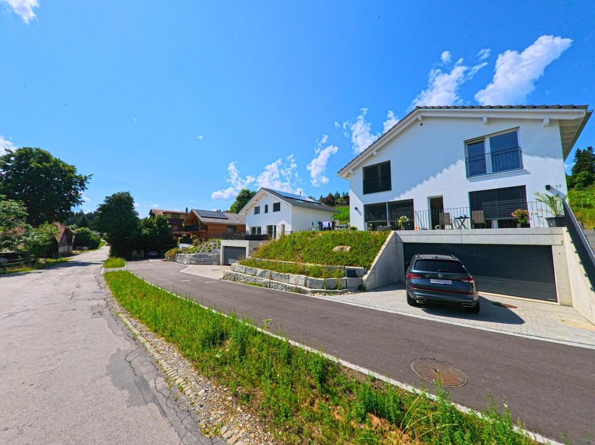 In Buchholterberg (3615, Kanton Bern) stehen moderne Einfamilienhaeuser mit Hanglage entlang einer asphaltierten Strasse, flankiert von Gruenstreifen und Baumbestand.