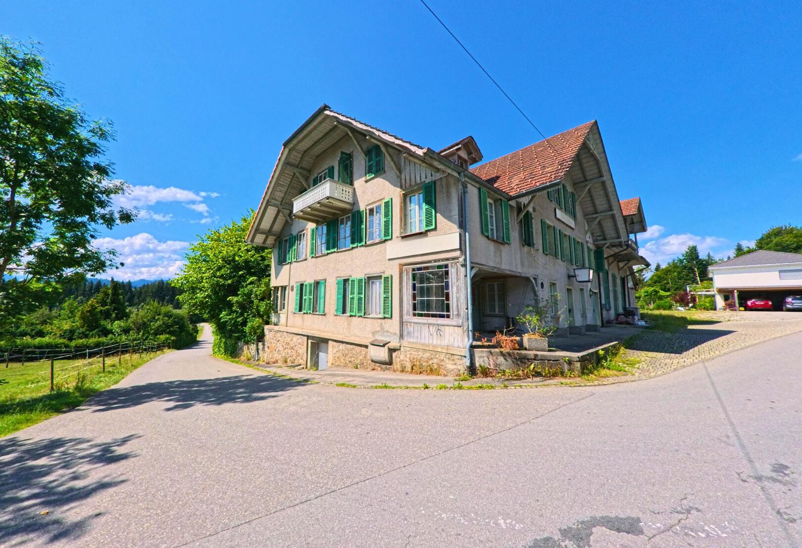 In Buchholterberg (3615, Kanton Bern) steht an einer Weggabelung ein traditionelles Wohnhaus mit gruenen Fensterlaeden und einem kleinen Balkon, umgeben von Baeumen und ländlicher Umgebung.