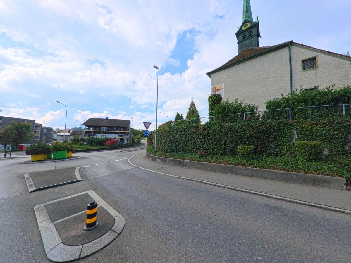 Eine Kirche mit gruenem Turm steht in Buchrain (6033, Kanton Luzern) an einer Strassenkreuzung mit bepflanzten Verkehrsinseln.