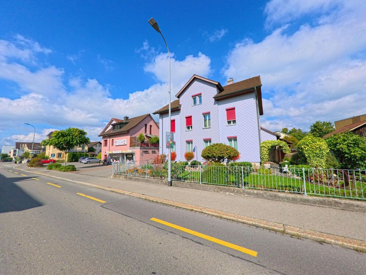 Entlang einer Strasse in Buchs (5033, Kanton Aargau) stehen gepflegte Wohnhäuser mit bunten Fassaden und blühenden Vorgärten im Sonnenschein.