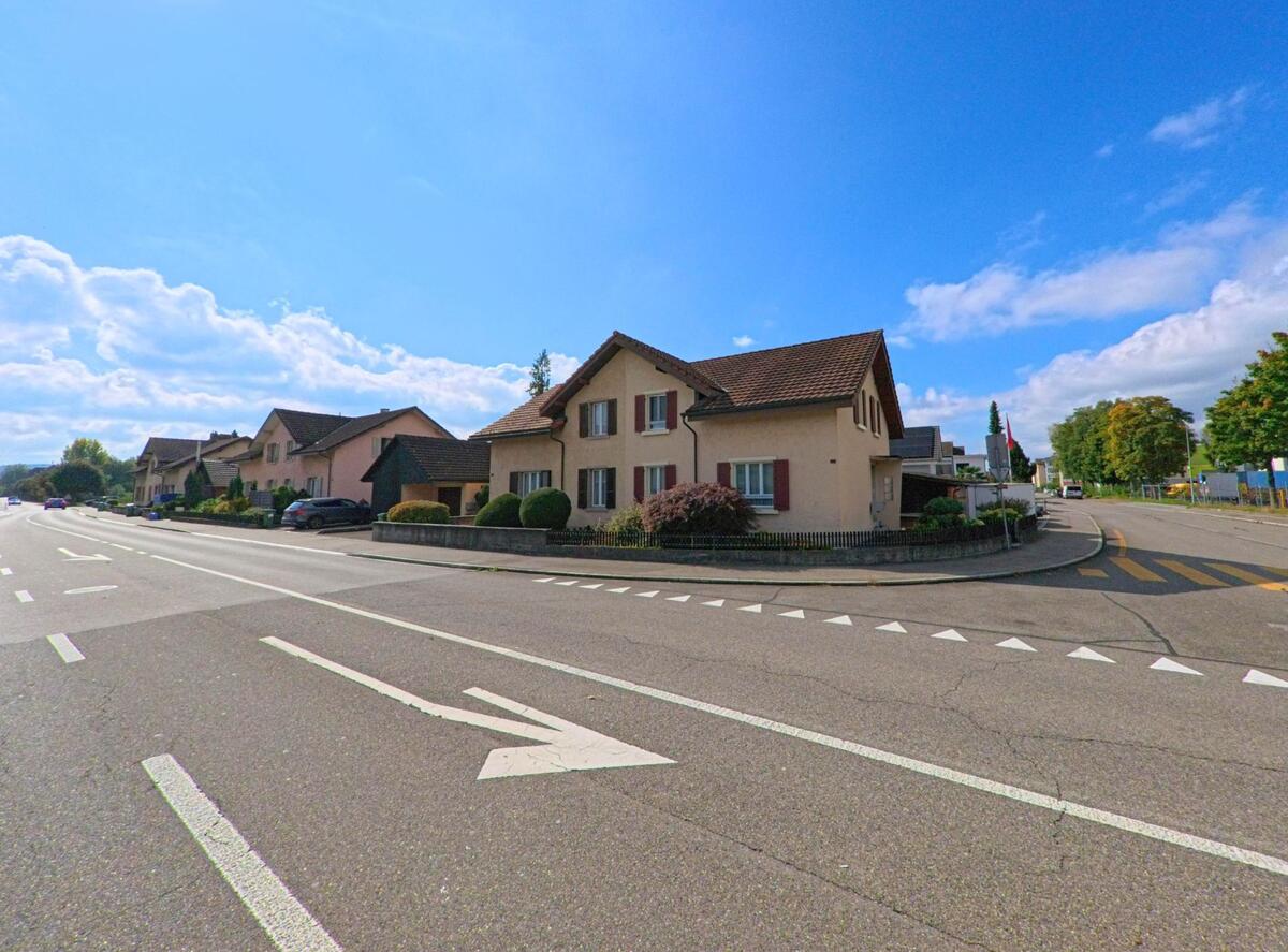 Entlang der Hauptstrasse in Buchs (5033, Kanton Aargau) stehen gepflegte Einfamilienhäuser mit Vorgärten, während der Verkehr über eine mehrspurige Fahrbahn führt.