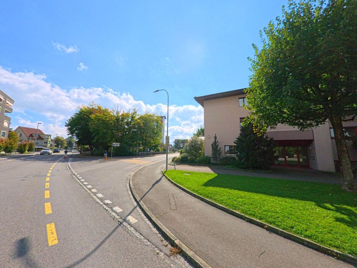 Eine breite Strasse mit Fussweg und Grünfläche führt in Buchs (5033, Kanton Aargau) vorbei an Wohnhäusern und schattenspendenden Bäumen.