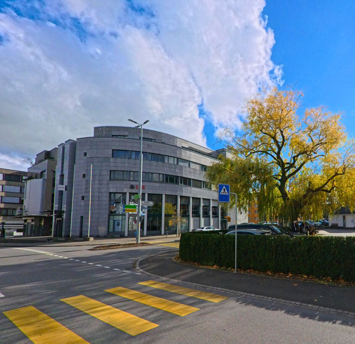Ein modernes Gebaeude mit abgerundeter Form praegt einen Strassenabschnitt in Buchs (9470, Kanton St. Gallen) nahe einem Fussgaengerstreifen.