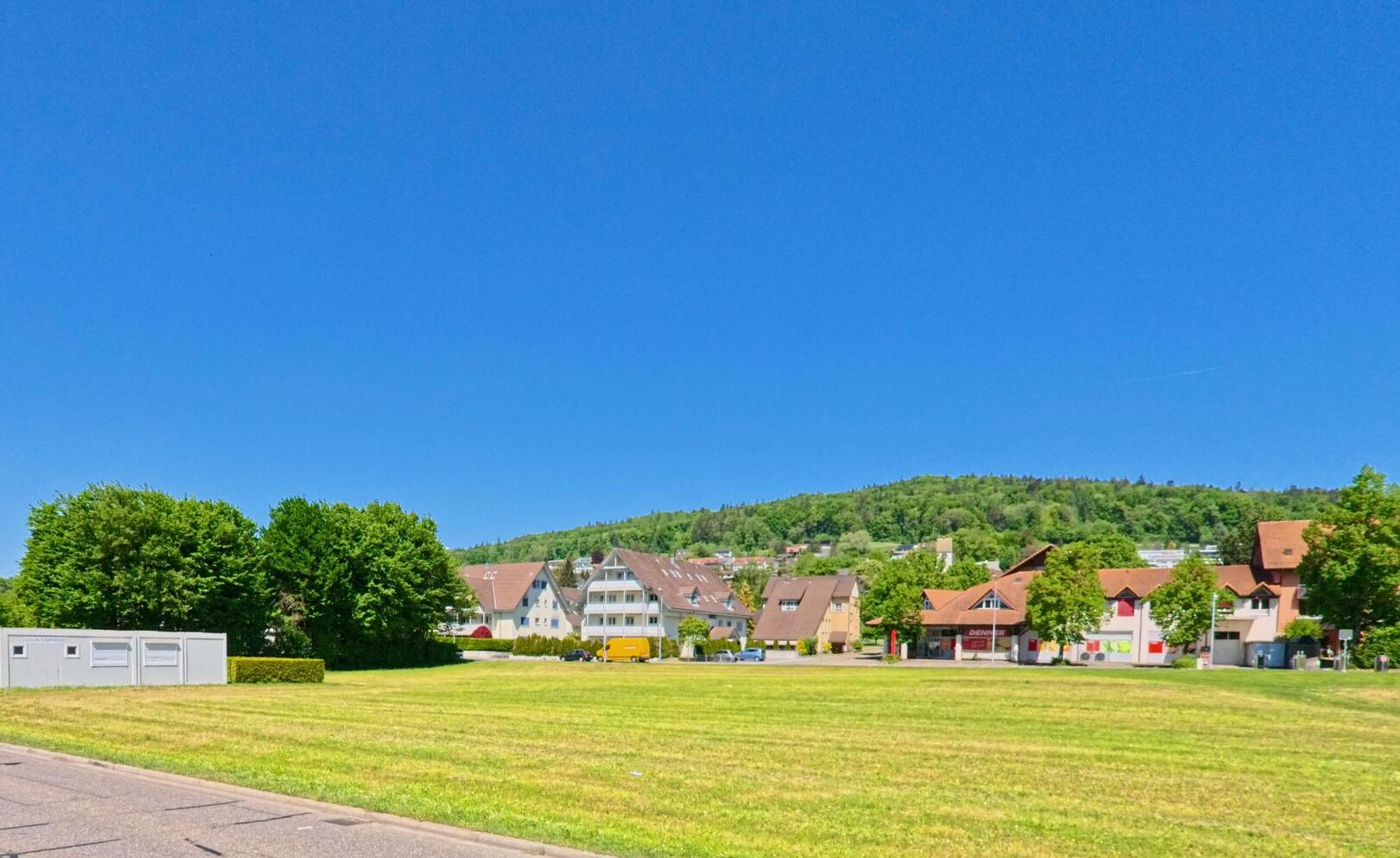 Vor der bewaldeten Hügellandschaft von Buchs im Kanton Zürich (PLZ 8107) liegt eine offene Wiese mit Blick auf Wohnhäuser und ein kleines Einkaufszentrum.