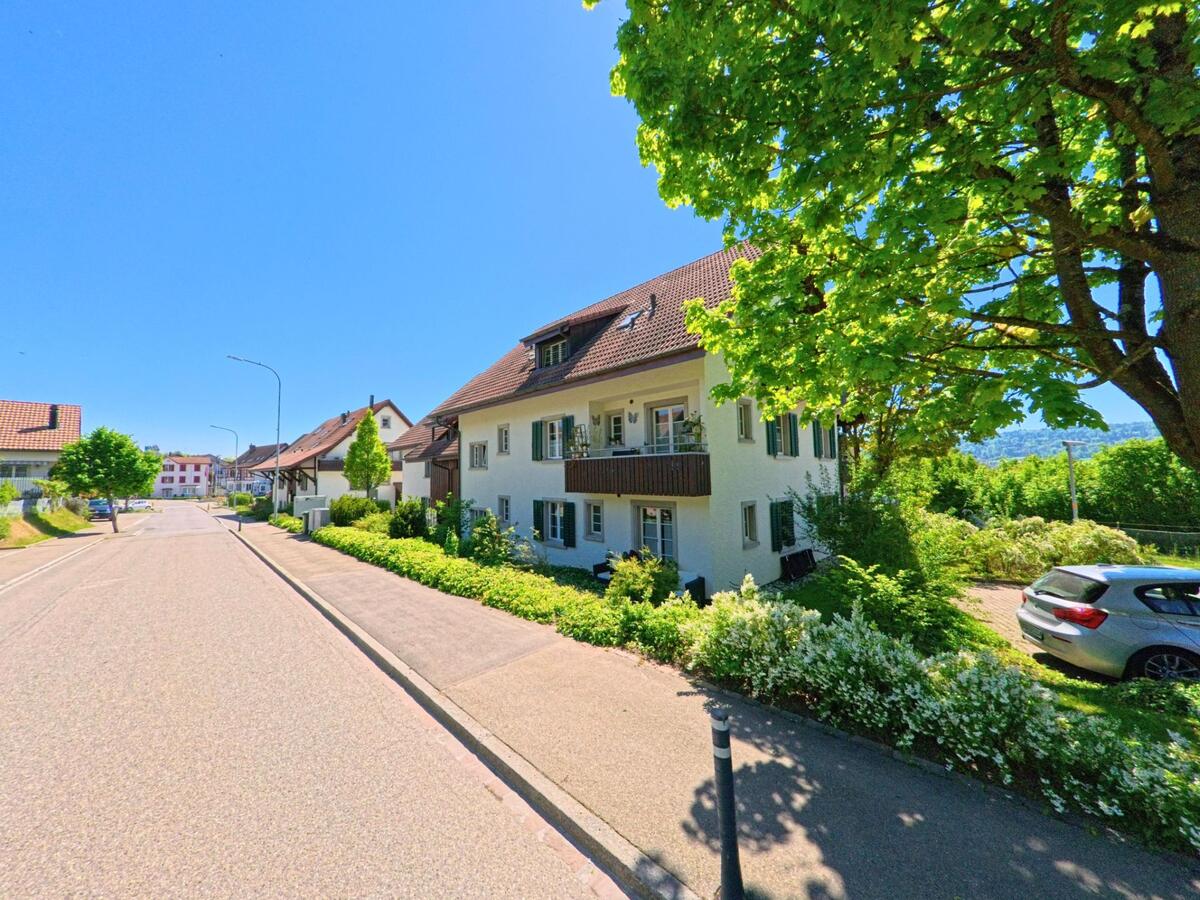 In Buchs im Kanton Zürich (PLZ 8107) säumen gepflegte Mehrfamilienhäuser mit Vorgärten eine ruhige Wohnstrasse unter schattenspendenden Bäumen.