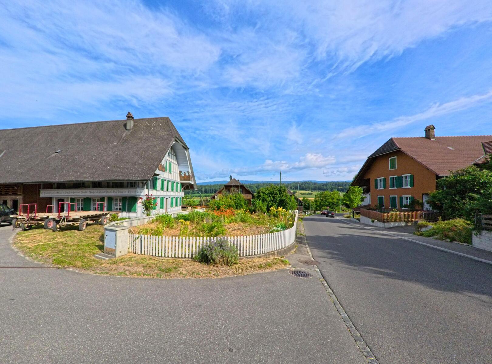 In Bühl (3274, Kanton Bern) prägt ein traditioneller Bauernhof mit grün-weissen Fensterläden das Dorfbild an einer ruhig gelegenen Kreuzung.