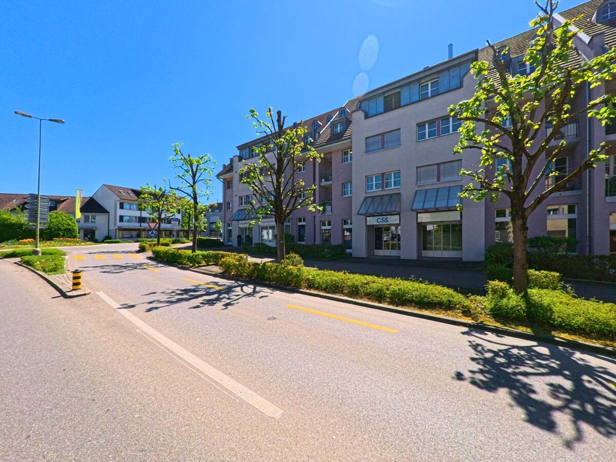 In Bülach im Kanton Zürich (PLZ 8180) säumen Geschäfts- und Wohngebäude mit Baumallee eine geteerte Strasse mit Mittelinsel und Zebrastreifen.