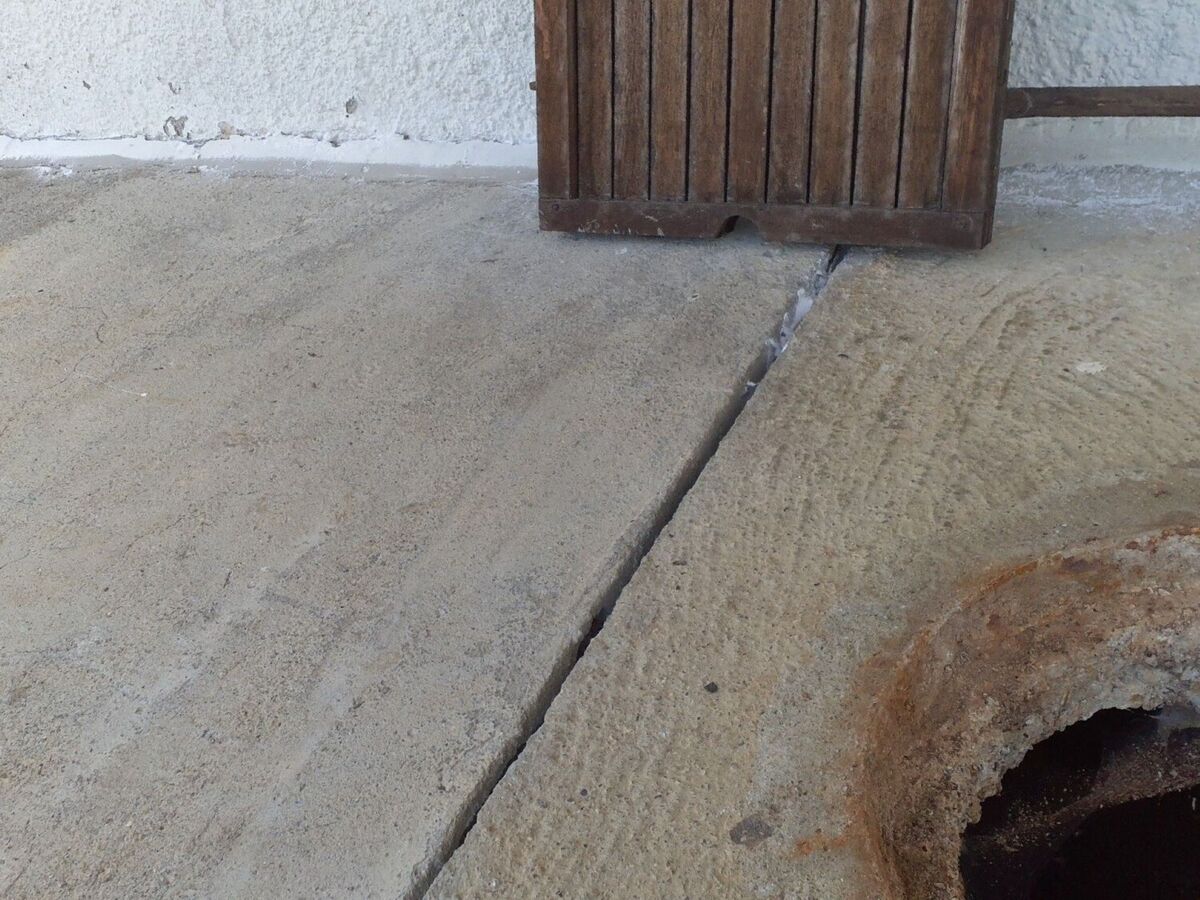 Eine Betonfuge neben einem verrosteten Gullydeckel zeigt in Bülach (8180, Kanton Zürich) feine Risse im Bodenbelag nahe einer weissen Hauswand.