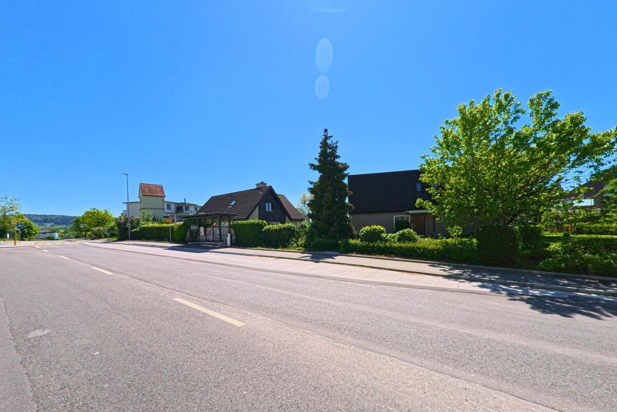 In Bülach im Kanton Zürich (PLZ 8180) liegt eine Bushaltestelle an einer breiten Strasse zwischen Einfamilienhäusern und gepflegten Vorgärten.